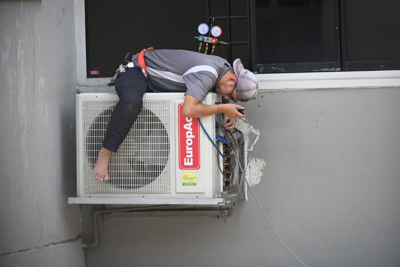 Technician working on a wall-mounted outdoor air conditioning unit with refrigerant gauges attached